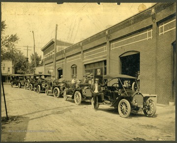 Cars and their owners are lined up in front of the brick Clarksburg Automobile Company building in Clarksburg, West Virginia.