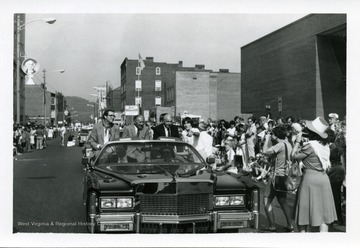 Jay Rockefller, an unidentified man, and Joe Dimaggio are riding in a car during a parade in the 1st Annual Italian Heritage Festival, Clarksburg, West Virginia. Tim Cotter (pointing)is standing next to the car.