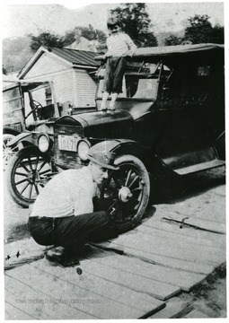 Earl Hugh Moneypenny crouched down near the front tire of his car. Small boy standing on car hood.  'Original photo owned by his daughter, Pearl Moneypenny of Clarksburg.'