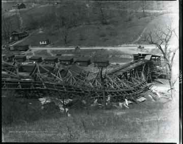 Coal mine, rail line and tipple overturned on a hillside.  A man stands among the wreckage and houses are visible along railroad tracks below.