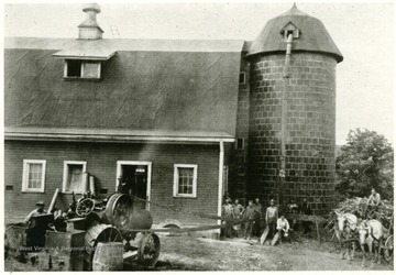 A belt is hooked up to a tractor. Group of men stand next to a silo attached to the barn.
