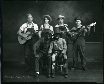 A group of entertainers hold their instruments and pose for a photograph.  Two seated are in blackface.