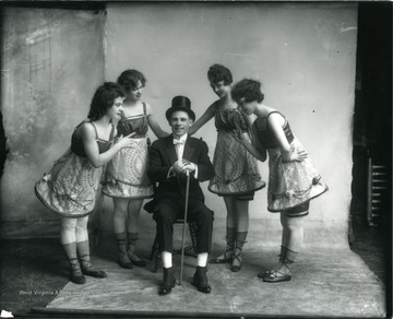 Four women performers standing around one male entertainer seated with a cane.