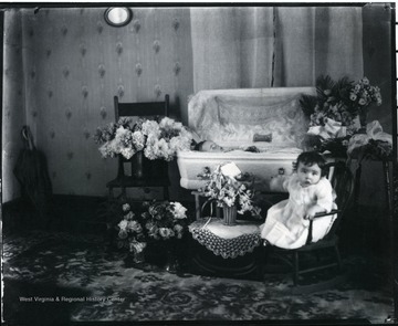 An unidentified child lays in a coffin while another child sits in a rocking chair near the casket.