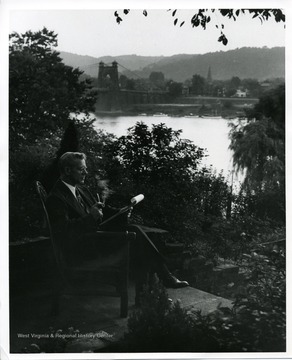 'The Old Timer' from the 'Its Wheeling Steel' radio program reads his latest script while relaxing near the Ohio river.  Wheeling Suspension bridge is visibl in the background.