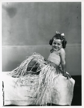 A smiling young girl is wearing a grass skirt while posing for the photographer.