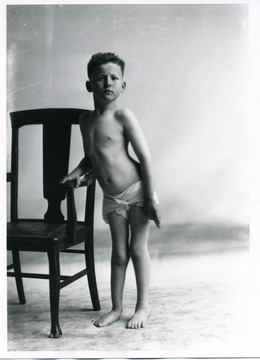 A young boy in a diaper standing next to a chair.