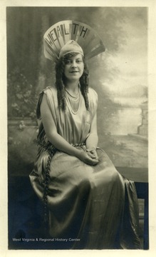 A picture postcard of a woman in costume, sitting in front of a painted background. She is wearing a hat with the word Wealth on it and a long dress. 