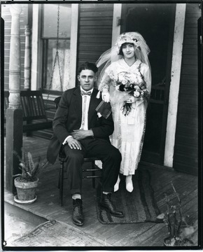 A bride stand behind the groom who is sitting in a chair on a front porch.