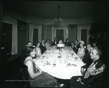 Group of people seated around a table with a wedding cake.