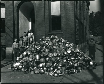 Group of people outside of Imperial Ice Cream Co. stand next to a large pile of tin buckets collected during a World War II Scrap Drive.  Imperial Ice Cream Company Manager, Roy Z. Burnside, standing on top step.