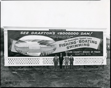 Four men stand in front of the billboard sponsored by the Taylor County Board of Trade advertising the Tygart Flood Control Dam and recreational activities on Tygart Lake.