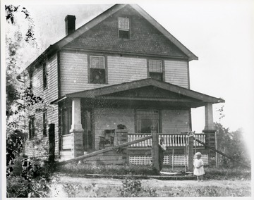 Two young children, an older boy and a younger girl, are standing by a gate near a porch of a two-story wooden house, location unknown.