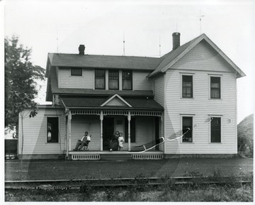 A man is sitting in a rocking chair while a lady is holding a child on the front porch, possibly West Virginia.