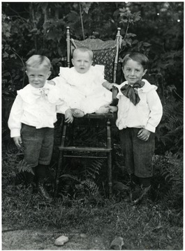 Two boys stand next to a baby sitting in a chair.