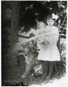 A young girl holding her doll is standing beside a dog.