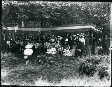 Large group of people gathered in the woods under a banner.  Many are holding American flags.