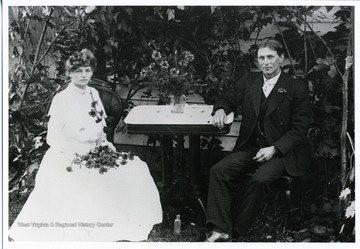Clara Koerner and John Gobeli are sitting at a table. Clara is wearing a long white dress and is holding flowers while John is wearing a three piece suit.  