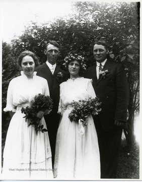 Two ladies and two gentlemen are standing together near a tree. The two ladies are holding flowers. Possibly a wedding portrait.