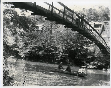 Three boys are standing along a wooden bridge looking down and watching four people enjoy a boat ride on an unidentified river in North Central, West Virginia.