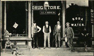 'Jack Crider's Drug Store, north side of 3rd Avenue between 10th and 11th Sts. Jake himself in the center of the door.  Man with the newspaper and a lean on the building is Cale C. Dusenberry.'