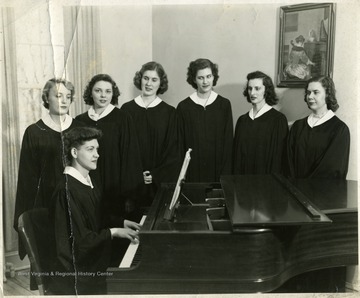 Six women standing and another woman sitting and playing the piano in choir robes.  Martinsburg, W. Va.