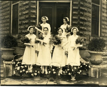 Six nurses standing on the stairs of the city hospital.  In their arms bouquets of flowers and in front of them baskets of flowers, Martinsburg, W. Va.