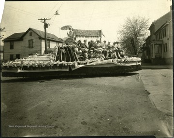 Group of kids and women on a float.  Woman holding an umbrella is seated on a barrel label Rising Sun Brand.