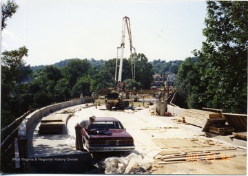 Unfinished bridge can be seen in progress. Workers surround equipment. 