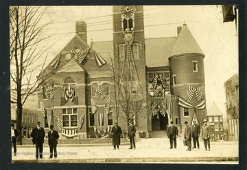 Several men are standing outside the Monongalia County Courthouse which is decorated with American Flags. The Courthouse is located on High Street between Pleasant and Walnut Streets in Morgantown, West Virginia.  Photo appeared in the Centennial Edition of the Dominion News.