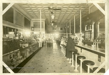 'This was a stationery and confectionery store, owned and operated by the Madeira Brothers, just below where Roger's Jewelry Store is now on High Street. They had the first soda fountain in town and were the first to bring the Pittsburgh papers to Morgantown. This picture was taken in the late 1880's. The gentleman seated is unidentified. At the fountain is B.H. Madeira and in the background Walter Madeira, uncle and father of Mrs. Lewis S. Core.' Centennial edition of Morgantown Post.