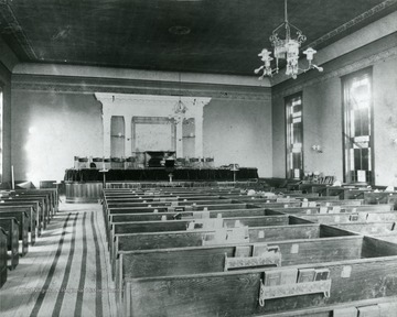 Interior of a Church located in Morgantown, W. Va.  Piano seen in pulpit. Chandeliers hang from the ceiling above pews. 