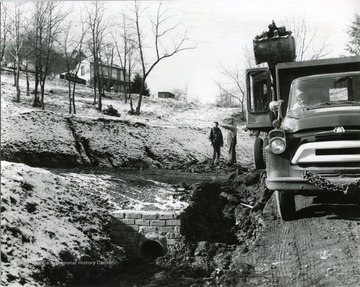 Men standing on road near culvert, talking with one another. Perhaps discussing construction plans for site. Trucks and equipment seen beside of workers. 