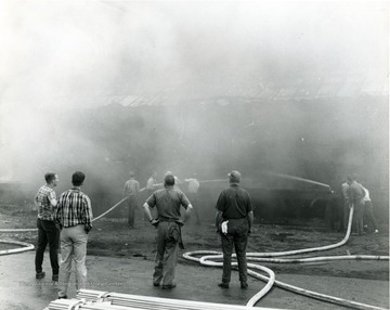 Firefighters work to extinguish fire at the Davis-Lynch Glass Company. Others stand amongst them watching the commotion. 