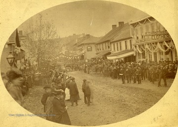 Parade on High Street in Morgantown, W. Va. Crowds flood the dirt-road. Horse-drawn carriages are seen amongst the crowd.  