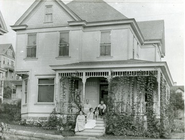 Home on Spruce Street in Morgantown, W. Va. People, possibly a family, sit on the front porch and steps with their dog. 
