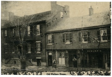 A man is standing on the stairs to the Old Wallace House in Morgantown, West Virginia. Two men are standing outside Dunlap's which is connected to the Old Wallace House.