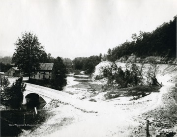 A bridge crosses a creek leading to a mill. Parts of the structure are missing. 