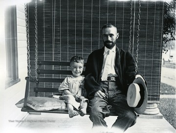 A man and a young child sit on this porch swing. Directly behind them is a hanging shade. 