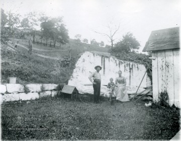 A family posing in a yard, near a large rock. On left side of family appears a dog house and on the right side of the family a shed. 