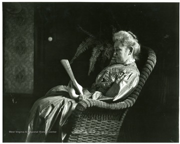 An elderly woman is sitting in a rocking chair reading a newspaper in a house in Morgantown, West Virginia.