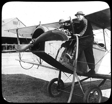 World War I Lantern Slide Show. In group of originally numbered slides.  (Number label is lost.)  Repair or maintenance of biplane engine on airfield.  There are two men standing adjacent to the fuselage on the wings.  A hanger containing an airplane can be seen in the background.  Frame is labelled with text saying 'Visual Bureau, University of Pittsburgh.'  (negative no. 23-531 is inscribed on slide)<br /><br />
