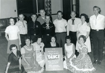 Group portrait of members of the Western Style Square Dance Club in Morgantown with a poster announcing their meeting time and place. 