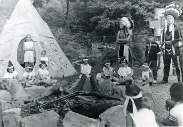 Group of children and adults dressed as Native Americans outside of a Teepee and around a campfire - possibly a 4-H group. 