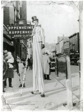 'Russell L. Long, Morgantown, W. Va. on high stilts, walks 3 to 4 hour periods on them.  This picture taken on High and corner of Pleasant Street - My construction of my stilts and the foot pivot is my own invention and makes it possible for me to move with comparative ease being a mechanical tall-man the hours I perform on them.  My leg stilts allow me to spread and stoop to go through doors from the sidewalks stilting indoors, and come out again on the sidewalk.  Many business places I have done stilting inside.'