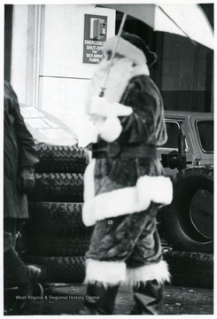 A man in a Santa Claus suit holds an umbrella at a service station.