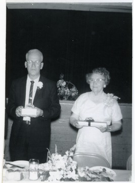An older couple stands holding their anniversary presents.