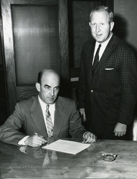 A portrait of Marlyn Lugar, sitting and Mal Campbell, standing. Mr. Lugar is signing a document while Mr. Campbell looks on.