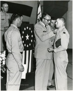 A man pins the collar of another man in uniform while others watch.