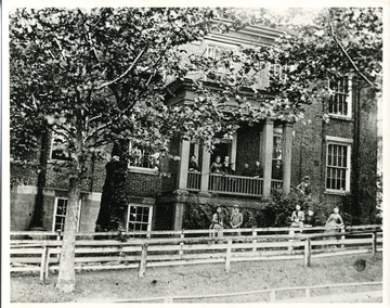 Women stand on the porch and in the lawn of the Morgantown Female Seminary on South High Street. The building burnt down in 1889.